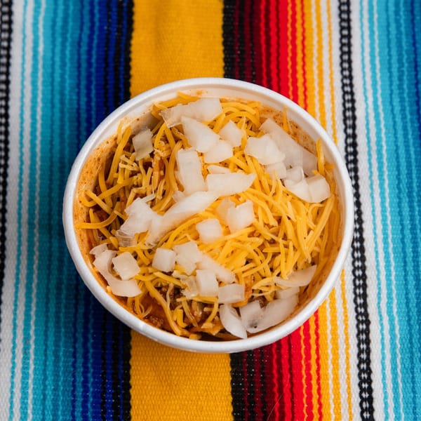 A white bowl of Pepe's Mexican Café chili with shredded cheese and diced onions on a colorful striped blanket.