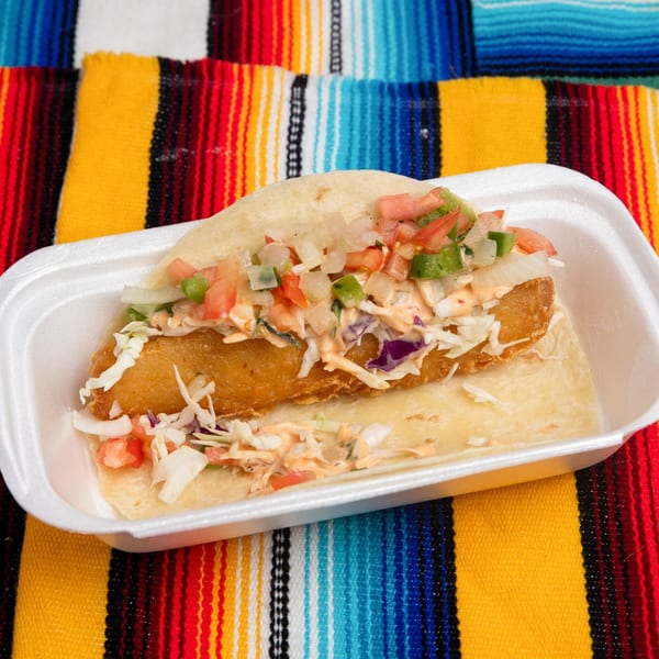 A Pepe's Mexican Café fish taco with pico de gallo and cabbage in a white container on a colorful serape.
